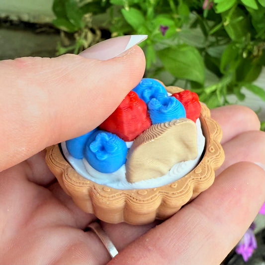 Hand holding a small wooden container with red, blue, and beige objects against a blurred green background