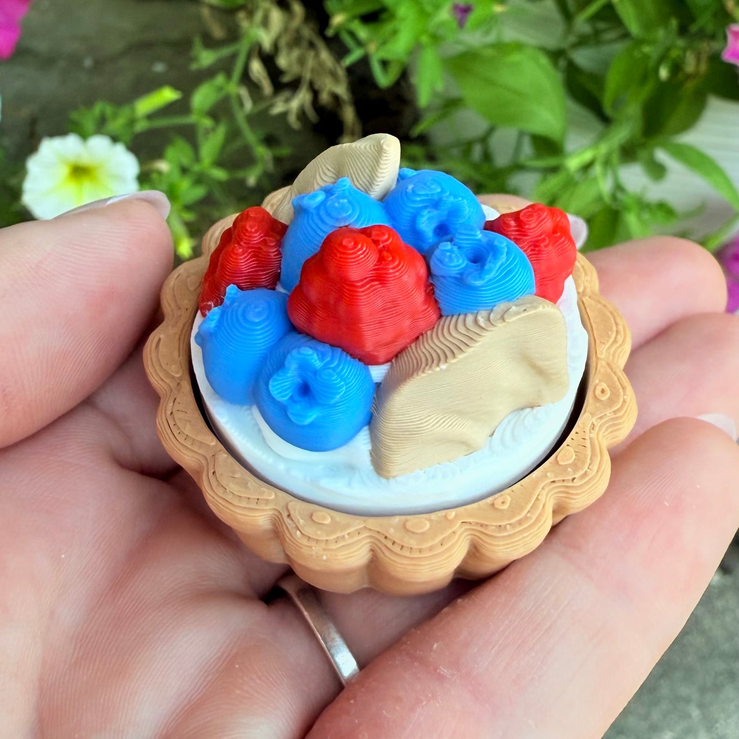 Hand holding a small decorative item with red, blue, and beige elements against a blurred natural background.